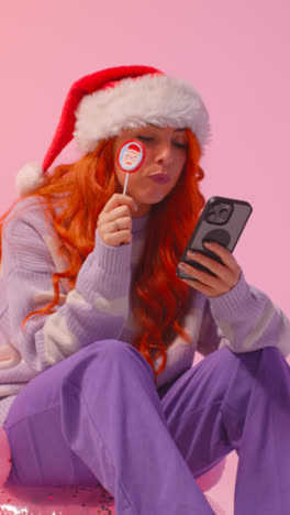 Vertical-Video-Studio-Shot-Of-Young-Gen-Z-Woman-Wearing-Christmas-Santa-Hat-Eating-Candy-Lollipop-Looking-At-Mobile-Phone